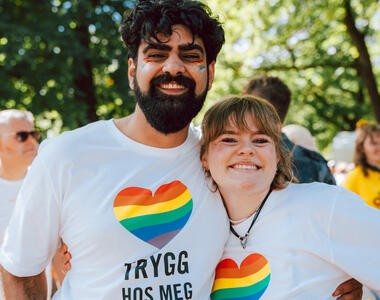 To personer smiler og holder rundt hverandre &ndash; begge har p&aring; t-skjorter med regnbuehjerte og teksten &laquo;trygg hos meg&raquo;.