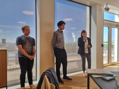 Tre mennesker som st&aring;r inntil en vegg med store vinduer. De presenterer